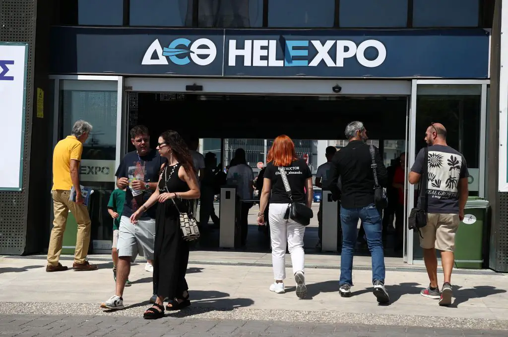 Unser Foto entstand vor dem Eingang des DETh-Messegeländes. Zu sehen ist das DETh-Helexpo-Logo. Darunter eine Eingangstür. Vier Personen sind dabei das Gebäude zu betreten, zwei haben es gerade verlassen.