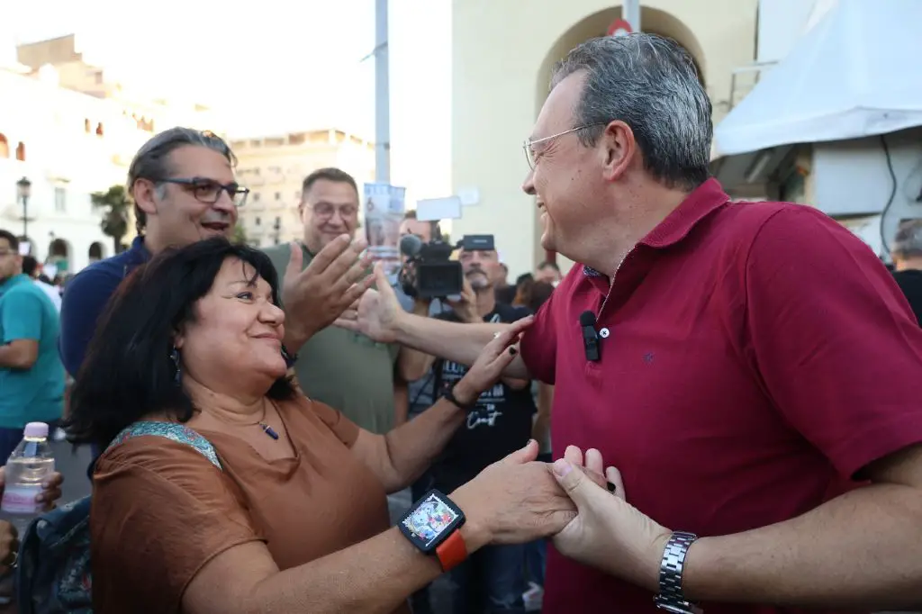 Unser Foto zeigt den Vorsitzenden der Oppositionspartei SYRIZA während einer Demonstration in Thessaloniki. Er trägt ein rotes T-Shirt. Mit Handschlägen begrüßt er eine Frau mit schulterlangen braunen Haaren und einen Mann, der neben ihr steht, und der ein blaues T-Shirt trägt.