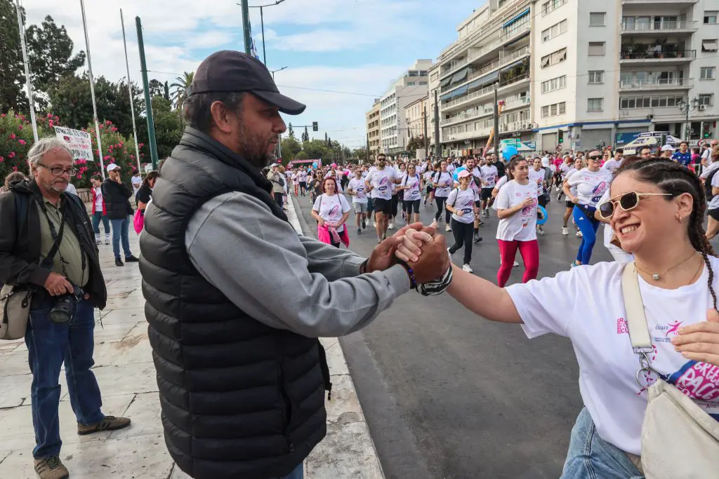 Unser Foto zeigt den hungerstreikenden Panos Routsis an der Amalias-Avenue vor dem Parlament. Er gibt einer vorbeijoggenden Frau die Hand, die ihre Haare in Zöpfe nach hinten geflochten hat. Sie zeigt ein breites Lächeln und trägt eine Sonnenbrille. Außerdem trägt sie ein weißes T-Shirt, auf dessen linker Seite vor rosa Hintergrund auf Englisch steht: „Greece Race for the Cure“. Hinter ihr joggen weitere Menschen – Frauen und Männer –, die das gleiche T-Shirt tragen. Auch Luftballons in rosa Farbe sind im Hintergrund zu sehen. 