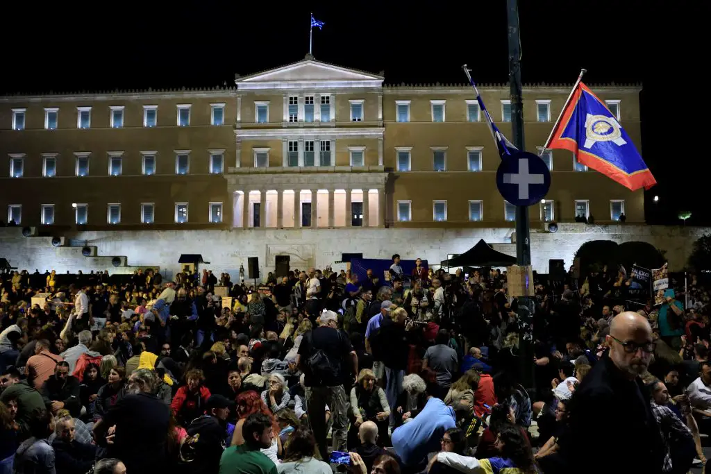 Unser Foto entstand in der Nacht. Es zeigt eine große Menschenmenge vor dem beleuchteten Parlamentsgebäude. Auf dem Dach des Parlaments ist eine griechische Flagge gehisst. An einem Mast auf dem Platz vor dem Parlament weht die Flagge der Stadt Athen neben der griechischen Fahne.  Viele Menschen sitzen auf dem Boden. Sie tragen langärmlige Kleidung.  Es dominieren die Farben schwarz, aber auch rot, blau und grün. Nur drei Personen tragen ein gelbes Oberteil. 