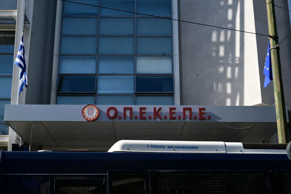 Unser Foto von Eurokinissi entstand vor OPEKEPE-Büros in Athen. Zu sehen sind die roten Buchstaben auf Griechisch: O.P.E.K.E.P.E. Dahinter sind eine blaue Wand und rechteckige Fenster, die mit Jalousien geschlossen sind. Davor sind links eine griechische Fahne und rechts eine Fahne der EU gehisst. 