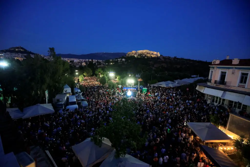 Unser Foto von Eurokinissi zeigt den Thission-Platz in Athen. Auf vier Straßen haben sich sehr viele Menschen versammelt, die dicht bei dicht nebeneinanderstehen. Am Straßenrand sind große weiße Sonnenschirme und weiße Mini-Vans von Kamerateams zu sehen. In der Mitte des Platzes ist eine beleuchtete Bühne aufgestellt. Dahinter sind Bäume und der Akropolis-Hügel sowie der beleuchtete Parthenon zu sehen. Dahinter zeigt sich ein Teil der Stadt und der Hymmetos-Berg. Der Himmel ist dunkelblau – er hat etwa die Farbe nach einem Sonnenuntergang.