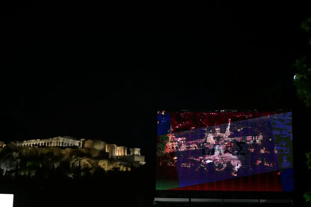 Dieses Foto von Eurokinissi entstand in der Nacht. Der Himmel ist fast schwarz. Das Foto zeigt einen Teil der beleuchteten Akropolis. Rechts ist ein Bildschirm, der den Thission-Platz abbildet, die Straßen sind voller Menschen.