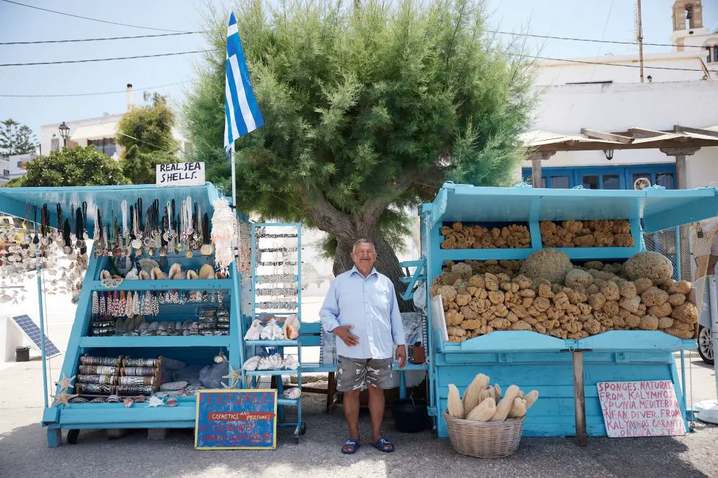 Unser Foto von Eurokinissi zeigt einen Verkäufer von Schwämmen aus der Insel Kalymnos. Er steht stolz zwischen seinem Stand mit Schwämmen und einem Stand mit Schmuck aus Muscheln. Er trägt ein hellblaues Hemd und eine kurze Hose. Hinter ihm ist ein Baum zu sehen.