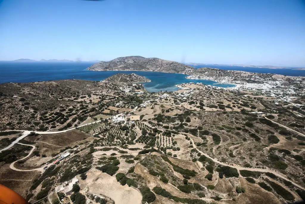 Unser Foto von Eurokinissi zeigt einen Teil der Insel Lipsi von Oben. Man sieht eine Buschlandschaft mit wenig Vegetation und ein Paar nicht asphaltierte Straßen. Im Bereich Mitte-Rechts des Bildes ist ein Ort mit weißen Häusern und Gebäuden und einem Hafen zu sehen. Die Farbe des Meeres im Hintergrund könnte man als Ägäis-Blau beschreiben.