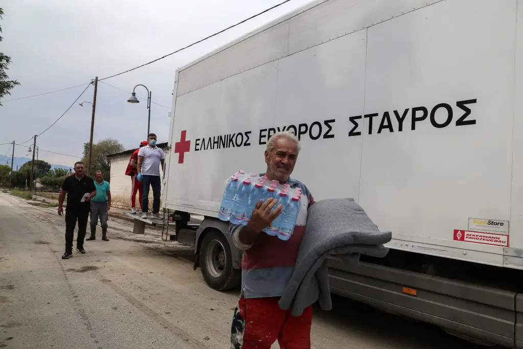 Im Foto ist ein Lastwagen des griechischen Roten Kreuzes zu sehen. Davor trägt ein Mann Wasserflaschen in der einen Hand und eine graue Decke in der anderen. Dahinter stehen Personen, die u. a. Handschuhe und Maske tragen.