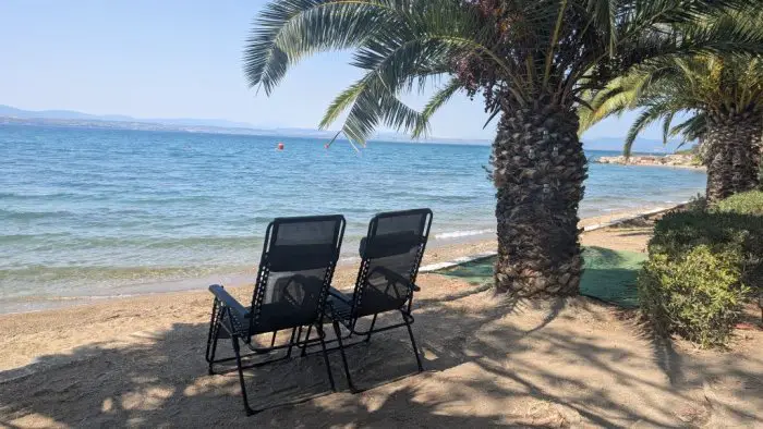 Zwei Liegestühle im Schatten einer Palme bieten Ruhe pur mit Blick aufs glitzernde Wasser (© Griechischenland Zeitung / Janrike Winter)
