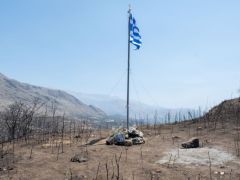 Unser Foto (© Eurokinissi) zeigt eine griechische Flagge in der verbrannten Landschaft südlich von Rethymnon, die an die beiden Feuerwehrleute Pantelis Diamantakis (25) und Manolis Stratidakis (58) erinnert, die während der Löscharbeiten ums Leben kamen.